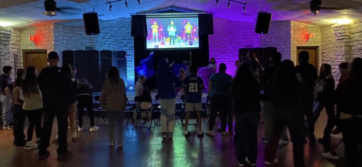 Children, teens, and adults worshipping together in a room with colorful lighting